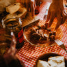 Plato de cristal típico de casa de pueblo donde están cortando Choricitos de orza picantes Arrrea, que han estado envasados en aceite de oliva. Al lado hay trozos de pan de pueblo cortados sobre una tabla. El mantel de la mesa sobre la que se apoya es típico de pueblo. Es la hora del aperitivo.